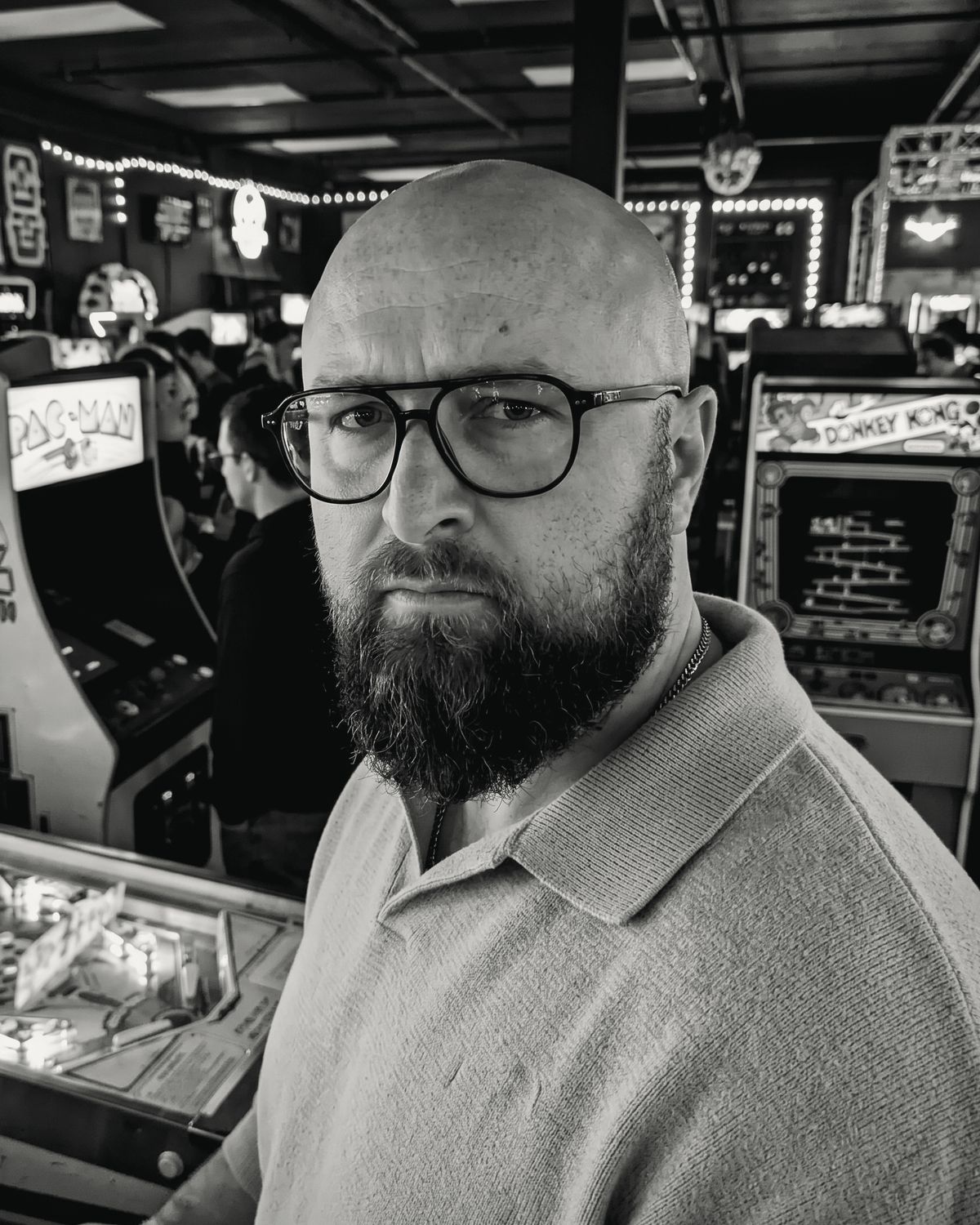 The principal of Liquid Lock, in black and white, standing in a vintage arcade between pinball and Donkey Kong cabinets
